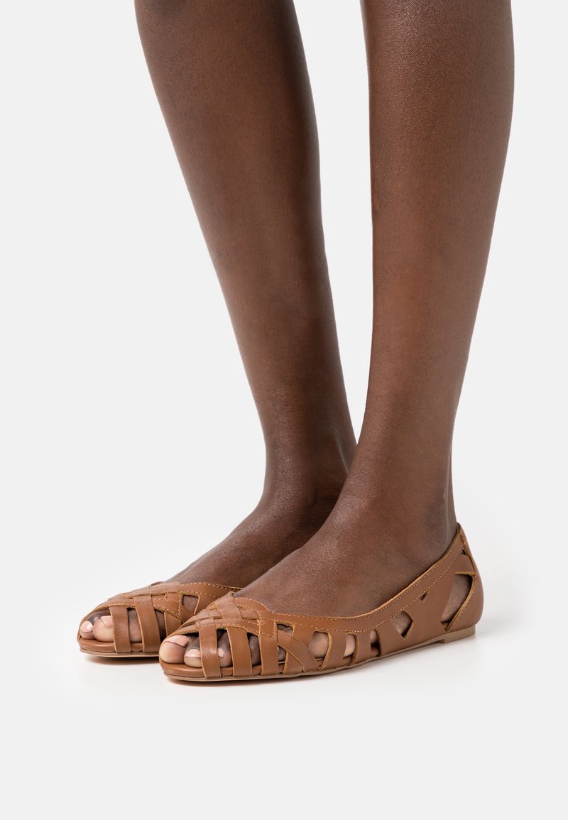 Chaussures plates en cuir tressé marron portées sur des pieds à la peau foncée sur un fond clair uni.
