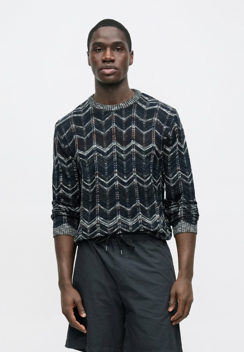Pull tricoté avec un motif en chevrons bleu marine et gris, doté d'un col rond côtelé et de manches longues, porté avec un short noir.