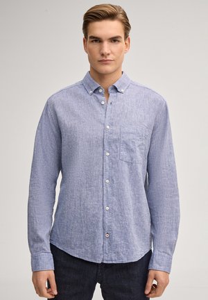 Junger Mann trägt ein hellblaues, knöpfbares Hemd mit Brusttasche und steht vor einem schlichten hellgrauen Hintergrund.