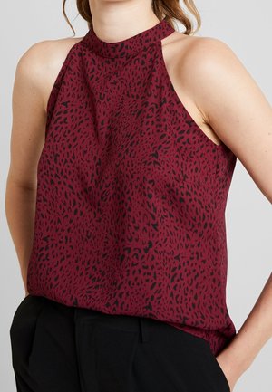 Sleeveless high-neck maroon top with black abstract animal print worn with black pants, hands in pockets.