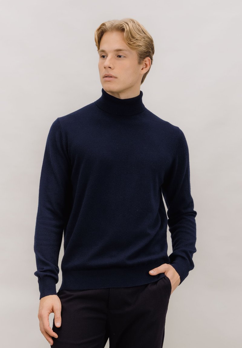 Marineblå genser med høy hals laget av mykt stoff, med lange ermer og ribbet nederkant, kombinert med mørke bukser. Jevn tekstur, minimalistisk design.