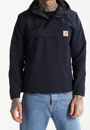 Mand iført en sort Carhartt hættejakke med frontlomme og blå jeans, synlige tatoverede hænder og nakke.