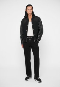 Hombre con cabello largo, vistiendo una chaqueta bomber negra, una camiseta sin mangas blanca, jeans negros y zapatillas negras, de pie contra un fondo blanco.