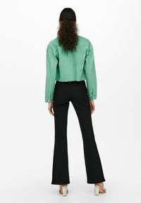 Giacca di jeans verde menta tagliata e pantaloni svasati neri indossati con sandali con tacco trasparente, visti da dietro su uno sfondo bianco.