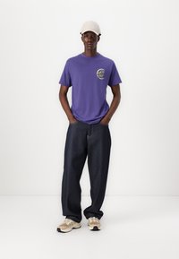 Purple cotton t-shirt with front logo, paired with loose dark denim jeans and beige sneakers. Model wearing a light beige cap.