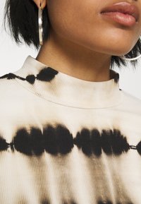 Jersey de cuello alto beige con canalé y un patrón tie-dye en negro. Presenta un cuello ajustado y tela texturizada. Visible un gran aro de plata.