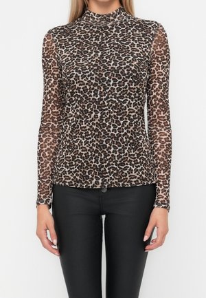 Vrouw met een langarmige top met hoge hals en luipaardprint, gecombineerd met zwarte aansluitende broek, staand voor een effen achtergrond.