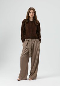 Bruine gebreide cardigan met ronde halslijn en knopen aan de voorkant, gecombineerd met een wijde beige broek met een gebonden ceintuur. Gladde textuur en ontspannen pasvorm.