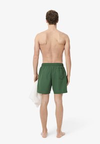 Shorts de bain verts avec une taille élastique et une poche arrière, portés par un homme tenant une serviette blanche, vus de dos.