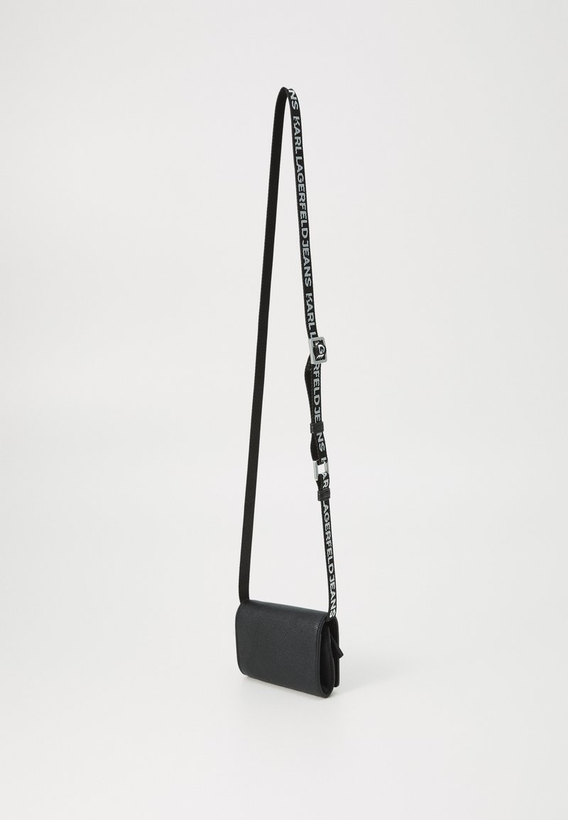 Sac bandoulière noir en matériau texturé. Présente une forme rectangulaire, une sangle réglable avec logo, et des accents en métal argenté.