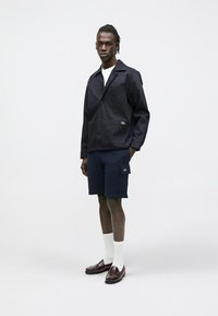 Tmavě modrá bavlněná bunda s náprsní kapsou a knoflíky, doplněná tmavě modrými šortkami s boční kapsou, bílými ponožkami a hnědými mokasínami.