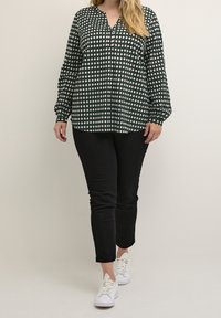 Groene blouse met lange mouwen en een wit ruitpatroon, met een V-hals en geknoopte mouwen, gecombineerd met zwarte slim-fit broek en witte sneakers.