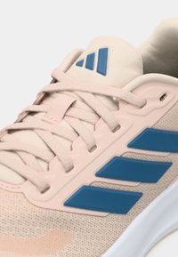 Gaiši rozā sporta apavi ar acs materiāla augšdaļu un zilu trīs svītru logotipu. Tajos ir plakanas šņorītes, teksturēti akcenti un bieza balta zole.