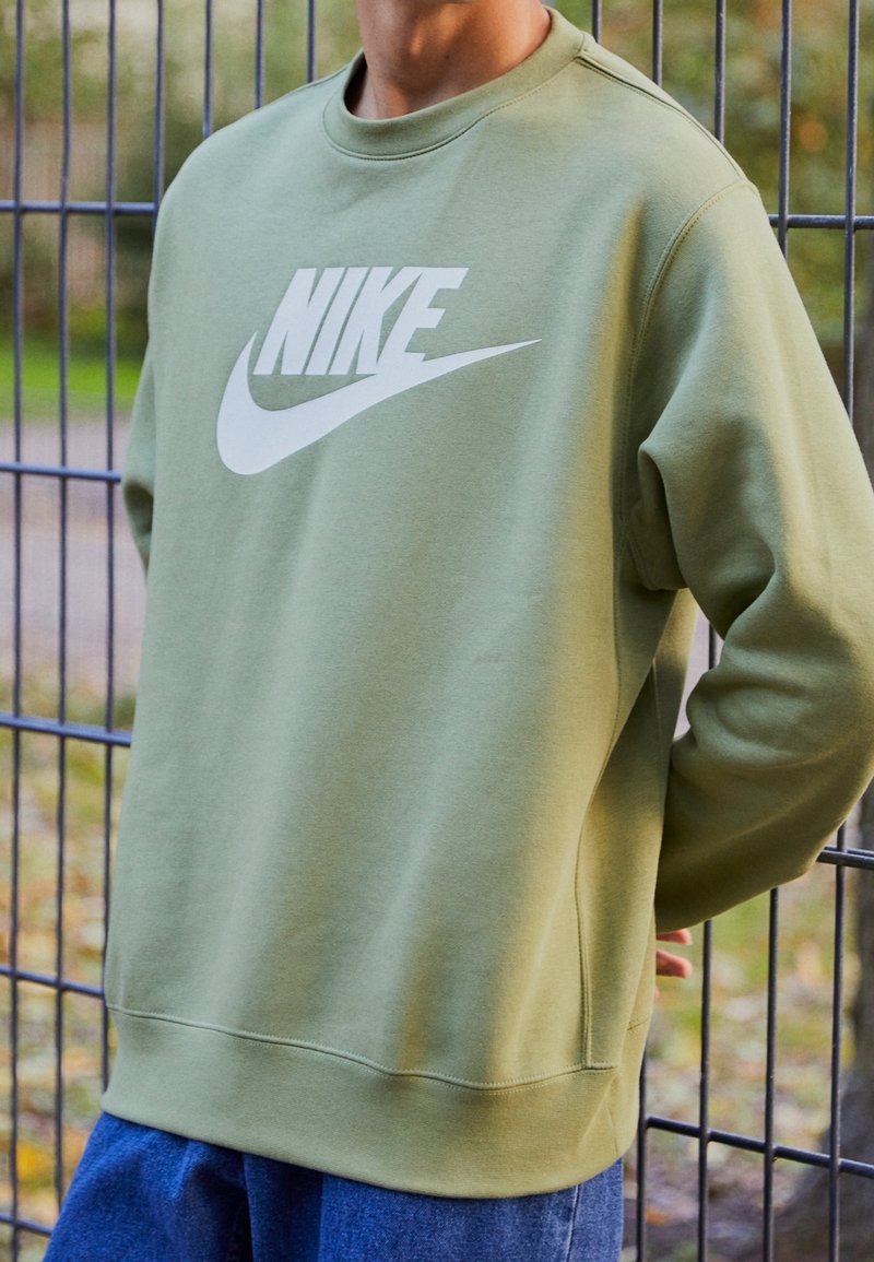 Olivgrön sweatshirt med en stor vit Nike-logotyp på framsidan. Har rund halsringning och ribbade ärmslut, placerad mot en stängselbakgrund.