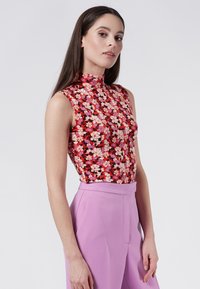 Top sin mangas de flores con cuello alto, con un fondo rojo y patrones florales en rosa, blanco y negro. Combinado con pantalones de talle alto en color lavanda.