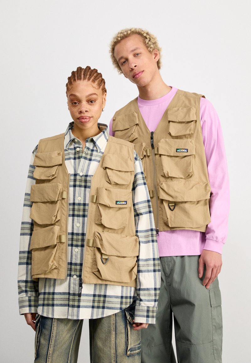 Beige Mehrtaschenwesten aus leichtem Material mit einem Reißverschluss auf der Vorderseite. Sie haben sechs externe Taschen und werden über einem karierten Hemd und einem hellrosa Oberteil getragen.
