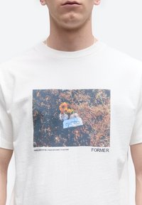 Vyras dėvi baltą marškinėlį su saulėgrąžų ir stiklainių nuotraukos spauda ant sauso žolės fono, apačioje užrašas „PASSING IMPERCEPTIBLY“ ir „FORMER“.