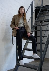 Chaqueta beige acolchada sobre una blusa verde claro, combinada con jeans negros y zapatillas blancas. De pie en unas escaleras de metal frente a una pared blanca.