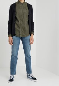 Granatowy kardigan na zielonej koszuli w kropki, jasnoniebieskie dżinsy i czarno-białe trampki Vans. Prosty design i casualowy krój.