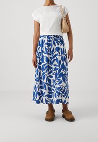 Valkoinen toppi, jossa on nyörityksellä tehty yksityiskohta, sinivalkoinen kukkakuvioinen hame, ruskeat puujalkineet, minimaliset asusteet. Rento kesäasu.