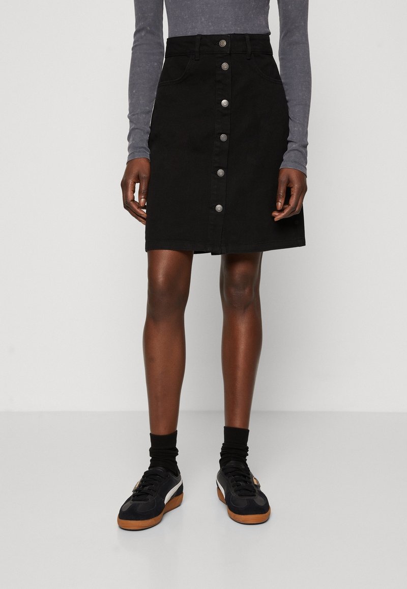 Musta nappisuljettav hame, jota on yhdistetty mustiin sukkiin, mustiin ja ruskeisiin lenkkikenkiin sekä harmaaseen pitkähihaiseen paita, jalat ja kädet näkyvissä.