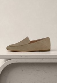 Ανοιχτό καφέ δερμάτινο μοκασίνι με ραμμένο επάνω μέρος, χαμηλό τακούνι και στρογγυλή μύτη, τοποθετημένο σε επιφάνεια με υφή πέτρας.