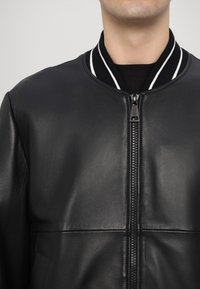 Jachetă bomber din piele neagră, cu guler din material ribat, având dungi albe, închidere cu fermoar frontal și textură netedă.