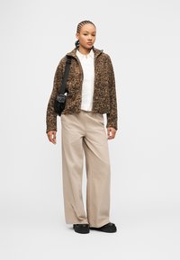 Νεαρή γυναίκα με πλεξούδες φοράει καφέ μπουφάν με φερμουάρ και σχέδια, λευκό πουκάμισο, μπεζ παντελόνι φαρδύ, μαύρα παπούτσια και μαύρη τσάντα χιαστί.