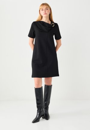 Vestito nero a maniche corte con scollo drappeggiato e dettaglio dorato, realizzato in un tessuto liscio. Abbinato a stivali aderenti neri fino al ginocchio.