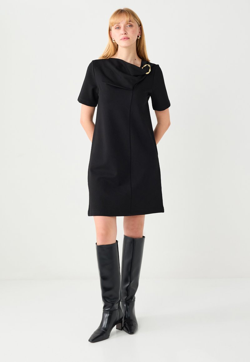 Robe noire à manches courtes avec encolure drapée et accent doré, en tissu lisse. Associée à des bottes noires ajustées montantes jusqu’au genou.