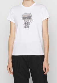 Valkoinen puuvillapaita, jossa on graafinen kuvasto hahmosta, joka näyttää aurinkolasit mustalla yksityiskohdalla, pyöreä pääntie ja lyhyet hihat.