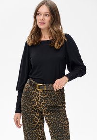 Kobieta z włosami do ramion w brązowym kolorze, ubrana w czarną bluzkę z długim rękawem oraz spodnie w leopardzi wzór z czarnym paskiem trzymającym klamrę.