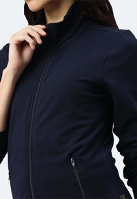 Giacca a zip blu navy realizzata in tessuto morbido, con colletto alto, tasche con zip e polsini a costine per una vestibilità aderente.