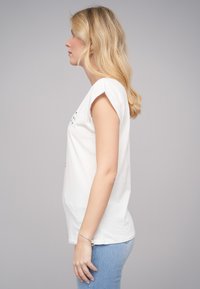 T-shirt en coton blanc avec des manches courtes roulées, présentant un design graphique sur la poitrine, associé à un jean en denim bleu clair.