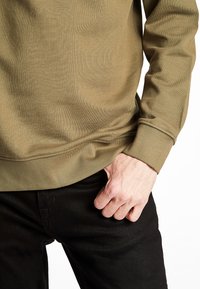 Felpa verde oliva con una texture liscia e polsini a costine, abbinata a jeans neri. Dettagli di cucitura evidenti lungo l'orlo.