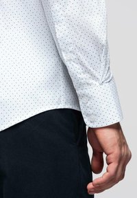 Main d'homme détendue sur le côté, portant une chemise blanche à petits points bleus et verts, et un pantalon bleu marine foncé.