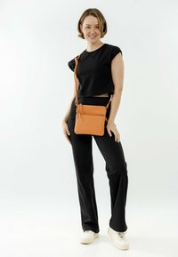 Oranžová crossbody taška vyrobená z texturované kůže s nastavitelným popruhem a zipovými kapsami, spárovaná s černým crop topem a černými kalhotami.