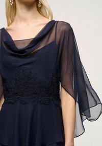 Mörkblå klänning med draperad halsringning, genomskinliga ärmar och intrikata spetsdetaljer vid midjan. Mjuk, flödande tyg med en strukturerad detalj.