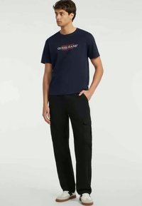 Nava majica iz bombaža slogom "GUESS JEANS" v beli in rdeči barvi. V kombinaciji z črnimi cargo hlačami in belimi supergami, ki imajo preprost dizajn.