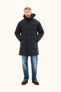 Wypchana granatowa kurtka z kapturem, z zapięciem na guziki i kieszeniami bocznymi, w połączeniu z jasnoniebieskimi dżinsami i brązowymi butami sznurowanymi.