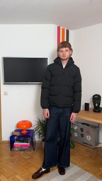 Melnā pūkainā jaka ar rāvējslēdzēju un kapuci, valkāta virs gaišas krekla, apvienota ar platajām tumšajām biksēm un spīdīgām brūnām kurpēm.