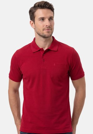 Mężczyzna ubrany w prostą, czerwoną koszulkę polo z krótkim rękawem, trzema guzikami i kieszenią na wysokości piersi, patrzący lekko w prawo na jednolitym tle.