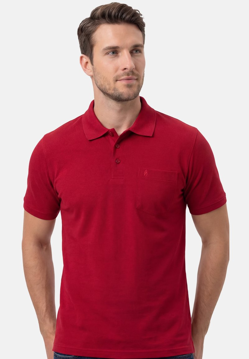 Homme portant un polo rouge uni à manches courtes avec trois boutons et une poche poitrine, regardant légèrement vers la droite sur un fond uni.