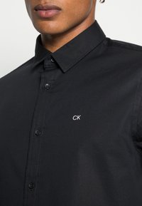 Camicia nera con colletto a punta, bottoni frontali e piccolo logo bianco "CK" sul lato sinistro. Tessuto liscio con dettagli minimi.