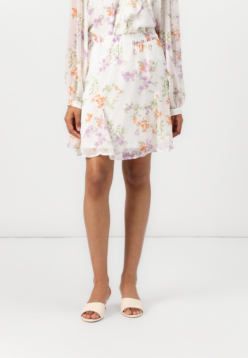 Jupe blanche à motifs floraux violet et orange, associée à un chemisier à manches longues assorti et des sandales beiges ouvertes à l'avant, portée par un mannequin.