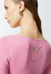 Ružičasti rebrasti pulover sa širokim izrezom, sa zlatnim grafičkim dizajnom na leđima. Model nosi zlatne naušnice u obliku obruča.