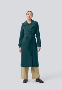 Woman wearing a long dark green belted coat, beige wide-leg pants, and black loafers, standing against a plain light background.
