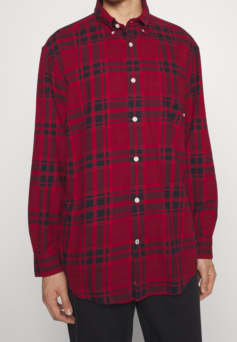 Chemise à carreaux rouge et noire à boutons, avec col, manches longues et une poche poitrine. Le tissu semble doux avec une finition texturée.