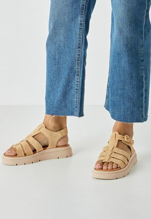 Sandales en daim beige avec plusieurs larges brides, une semelle épaisse et un détail à boucle, portées avec un jean denim raccourci.