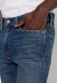 Main à la peau foncée partiellement dans la poche avant d'un jean bleu porté avec une chemise blanche.